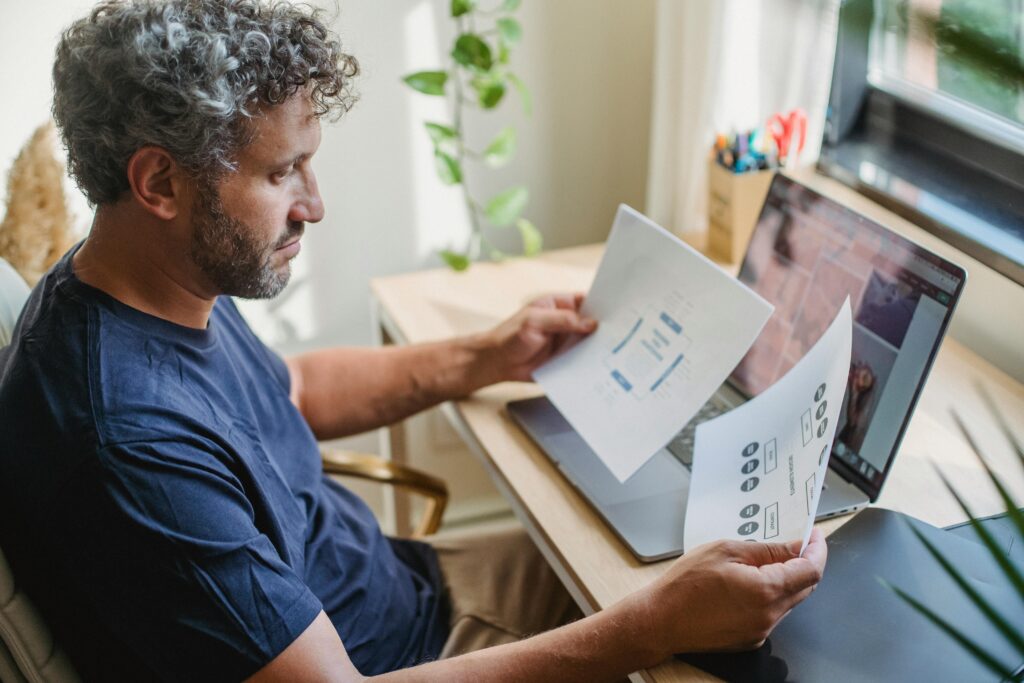 Focused adult male with beard and gray curly hair in casual clothes sitting at table with laptop open while working with papers